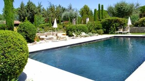 Una piscina rectangular con tumbonas y sombrillas, rodeada de setos y árboles cuidadosamente recortados.
