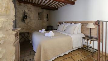 Un dormitorio con cabecera de madera, cama con ropa de cama blanca, mesita de noche con lámpara y pared de piedra.