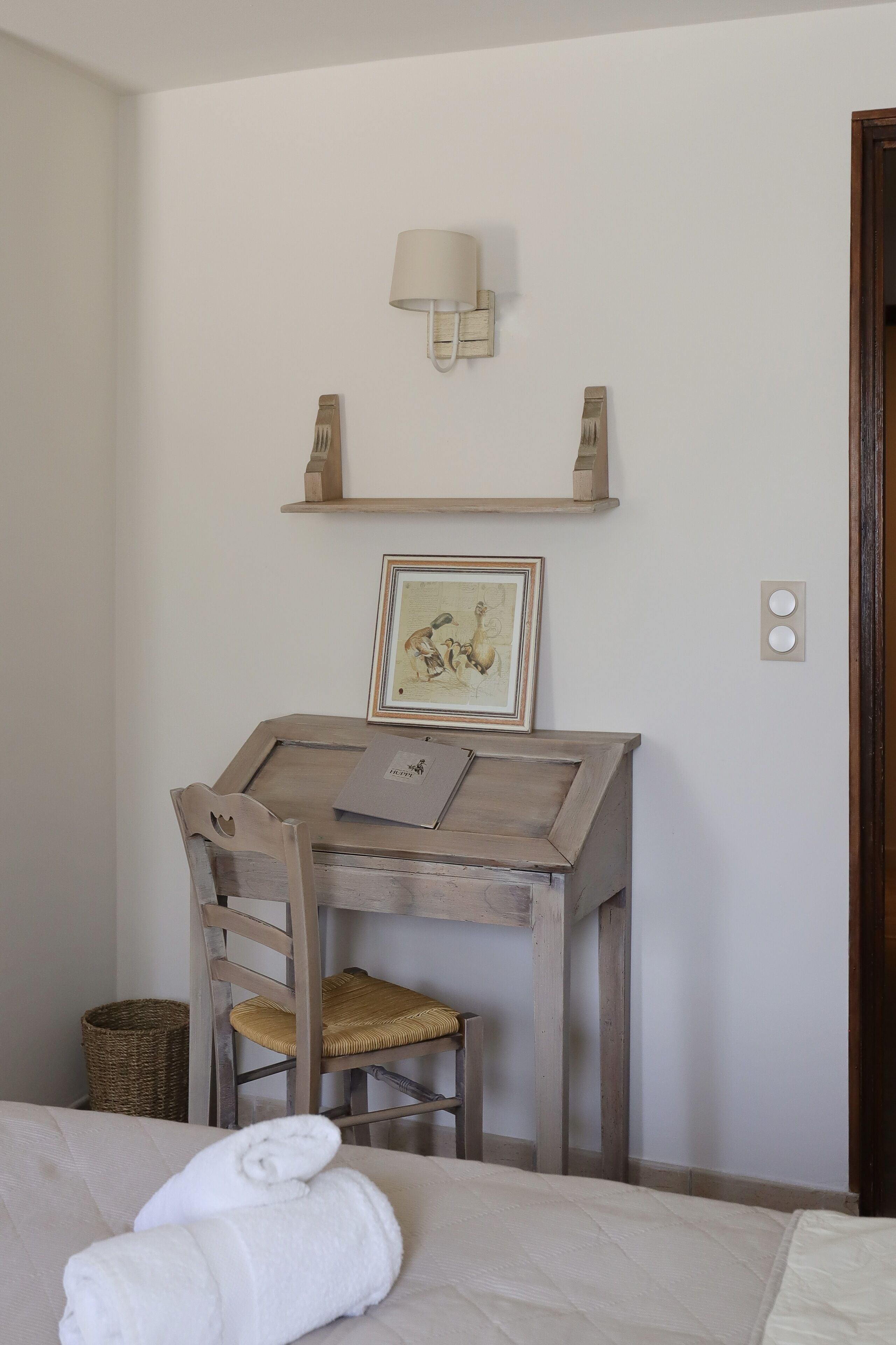 A bedroom with a bed, a wooden desk with a chair, a picture frame, a lamp, and a wall-mounted shelf.
