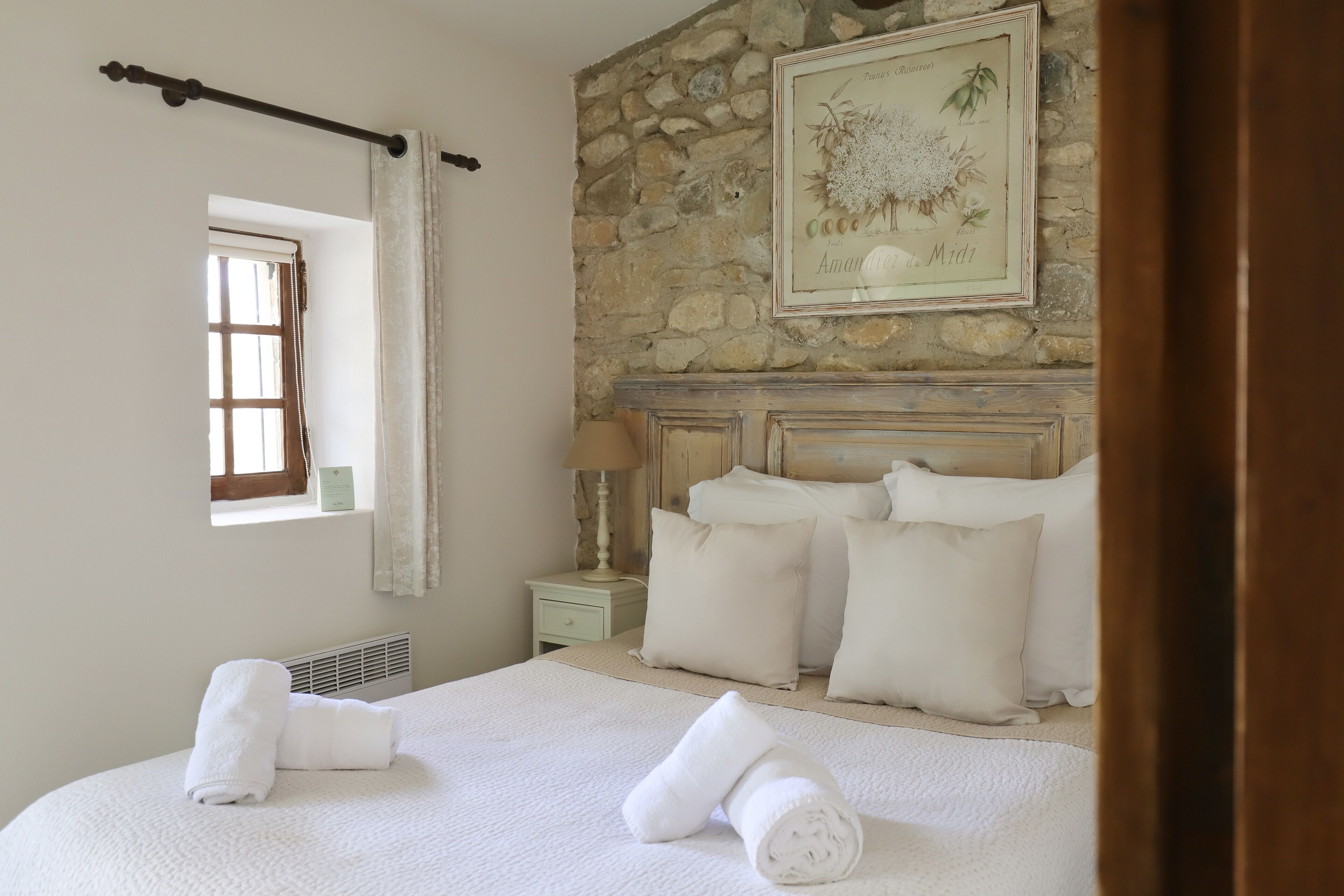 Un dormitorio con pared de piedra, cama con ropa de cama blanca, mesita de noche con lámpara y ventana con cortinas.