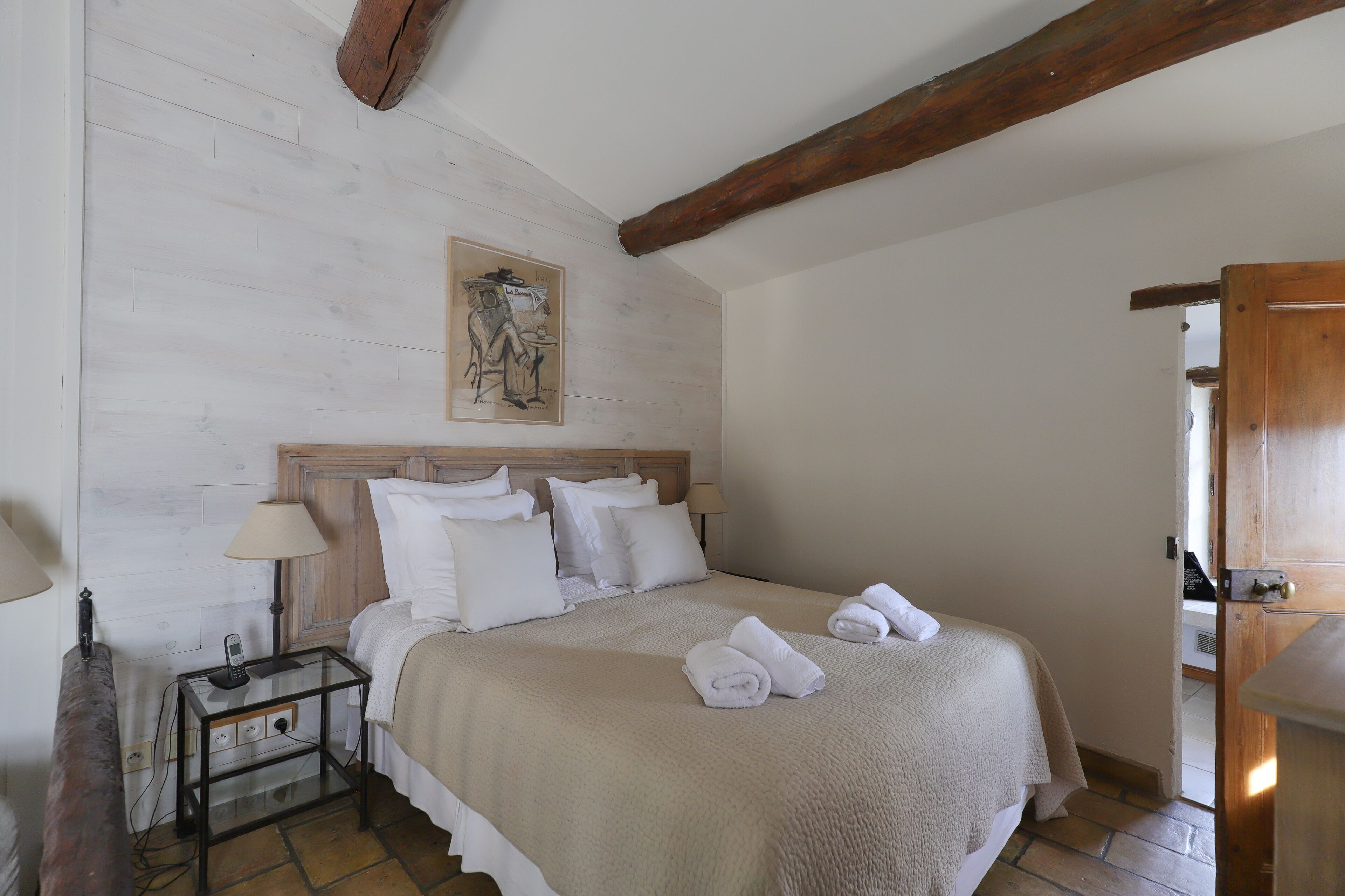 Een slaapkamer met een houten hoofdbord, een bed met witte lakens, een nachtkastje met een lamp en een schilderij aan de muur.