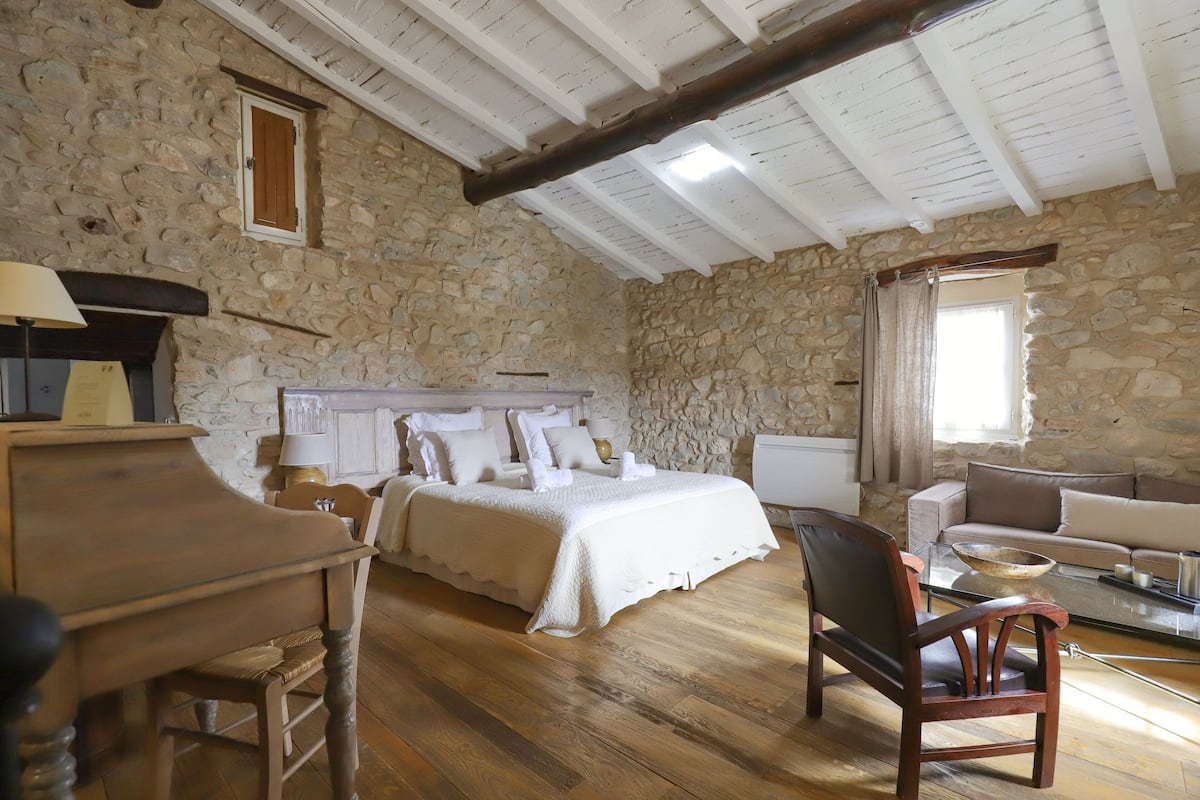 Une pièce avec un mur en pierre, des poutres apparentes en bois au plafond, un lit avec une literie blanche, un bureau en bois, une chaise et un canapé.