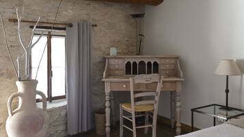A bedroom with a wooden desk, a chair, a bedside table, a lamp, and a vase with branches.