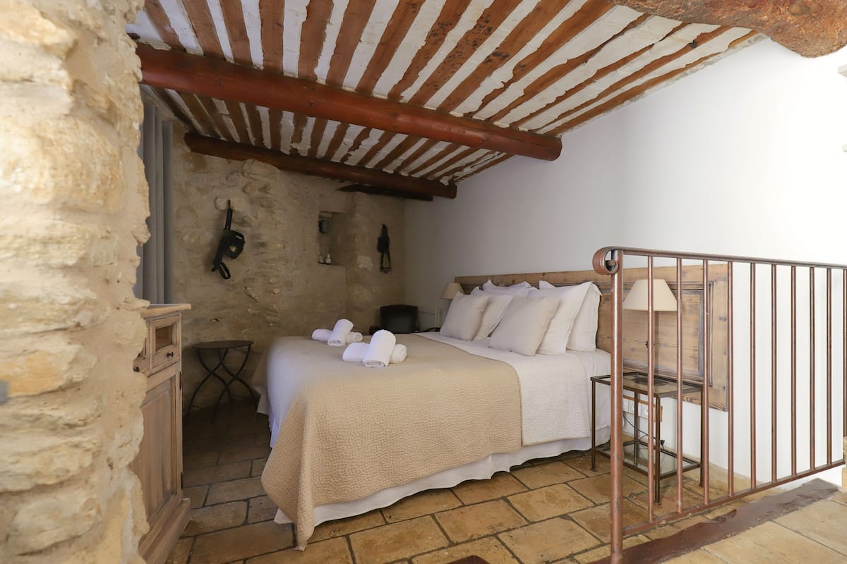 Une chambre avec un mur en pierre, un plafond en bois, un lit avec du linge de lit blanc et une petite table avec une lampe.