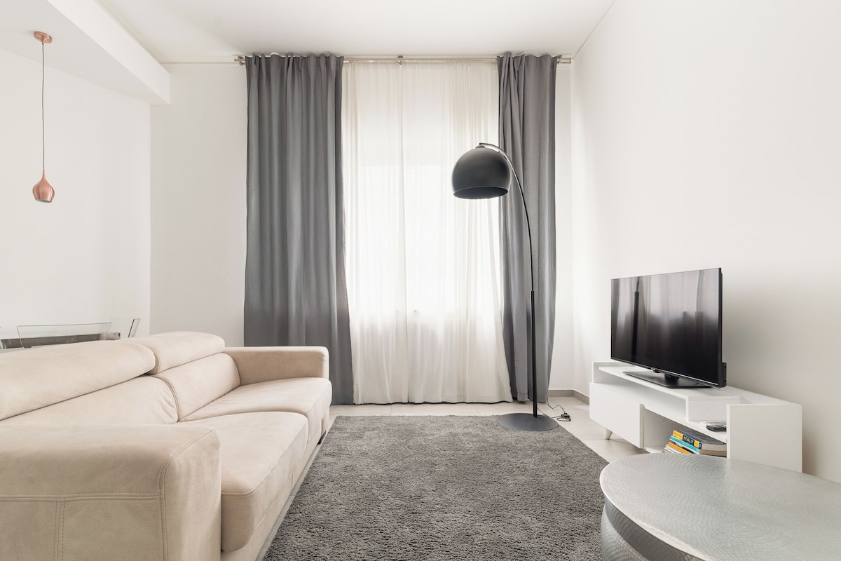 A modern living room with a sofa, a flat-screen TV, a floor lamp, and a large window with curtains.