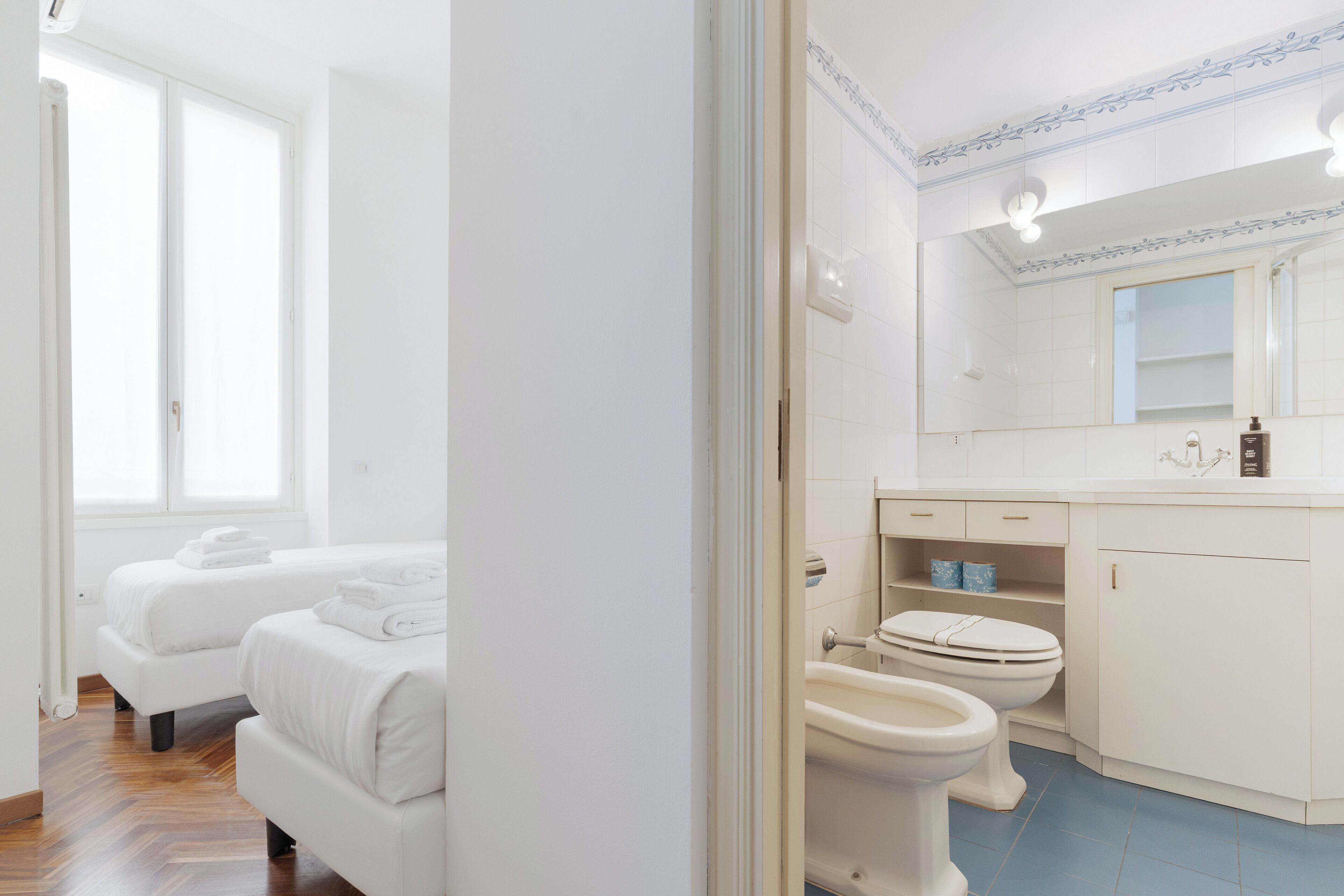 A modern bathroom with a toilet, bidet, and a large mirror. There are two white beds in the adjacent room.