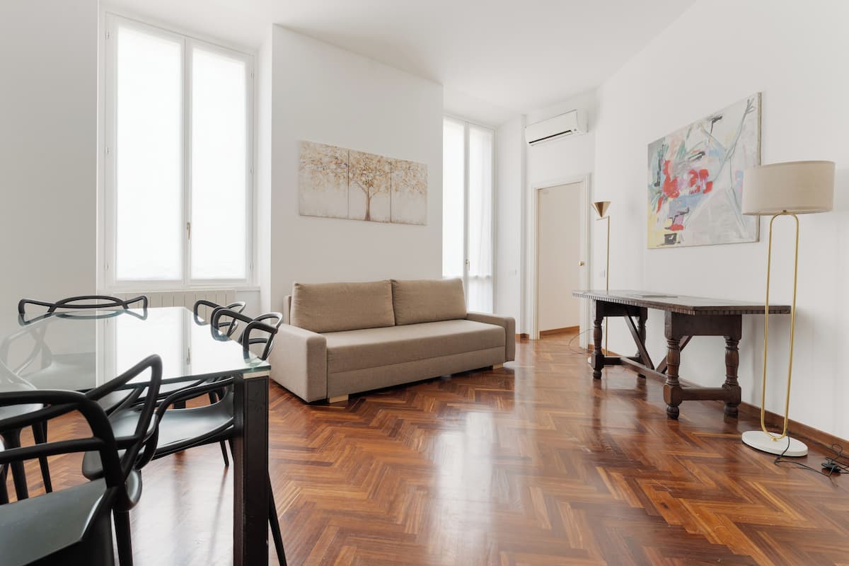 A modern living room with a glass dining table, a beige sofa, and a wooden floor.