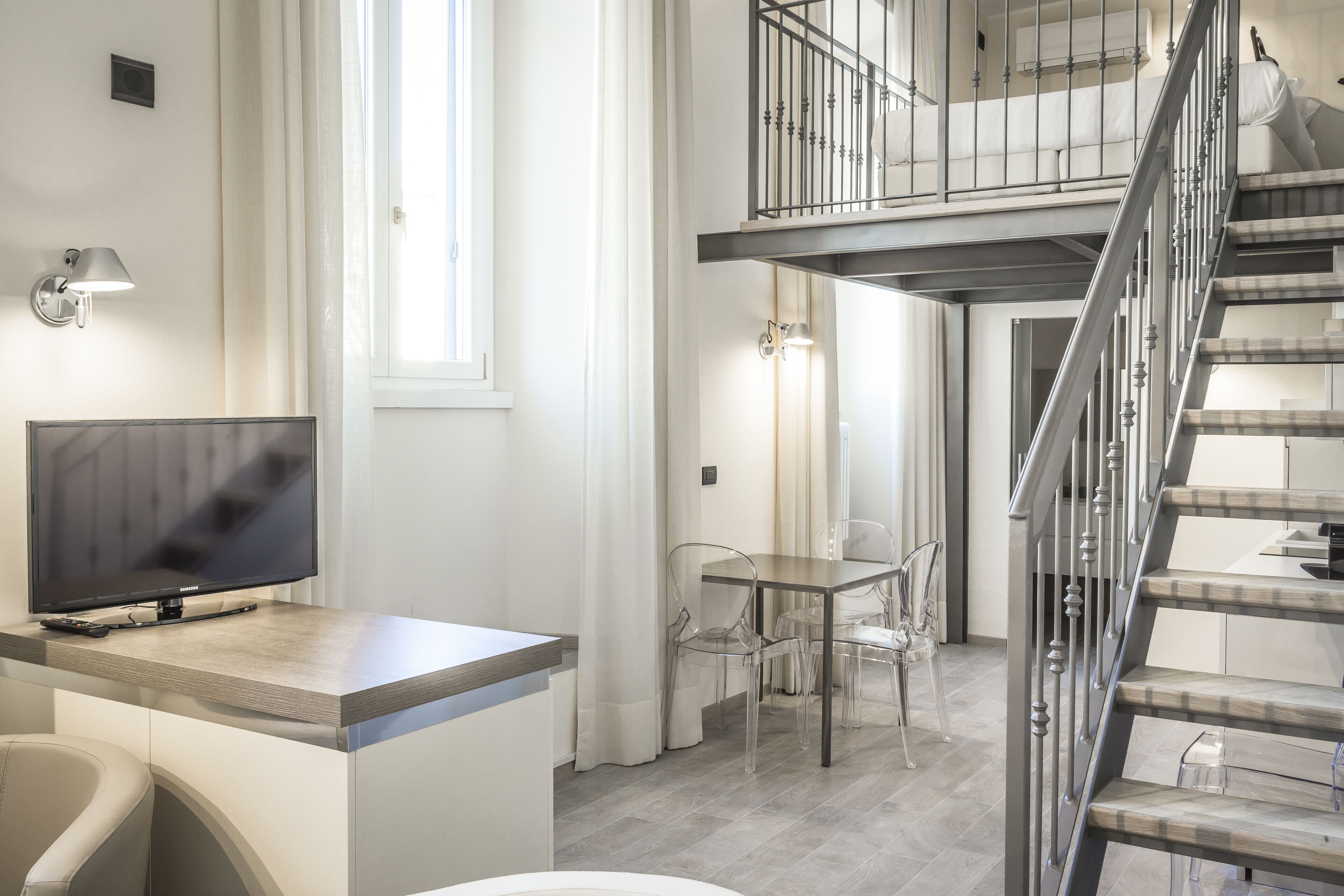 Un interior moderno con un televisor de pantalla plana sobre un escritorio de madera, una zona de comedor con sillas traslúcidas y una escalera con barandilla de metal.