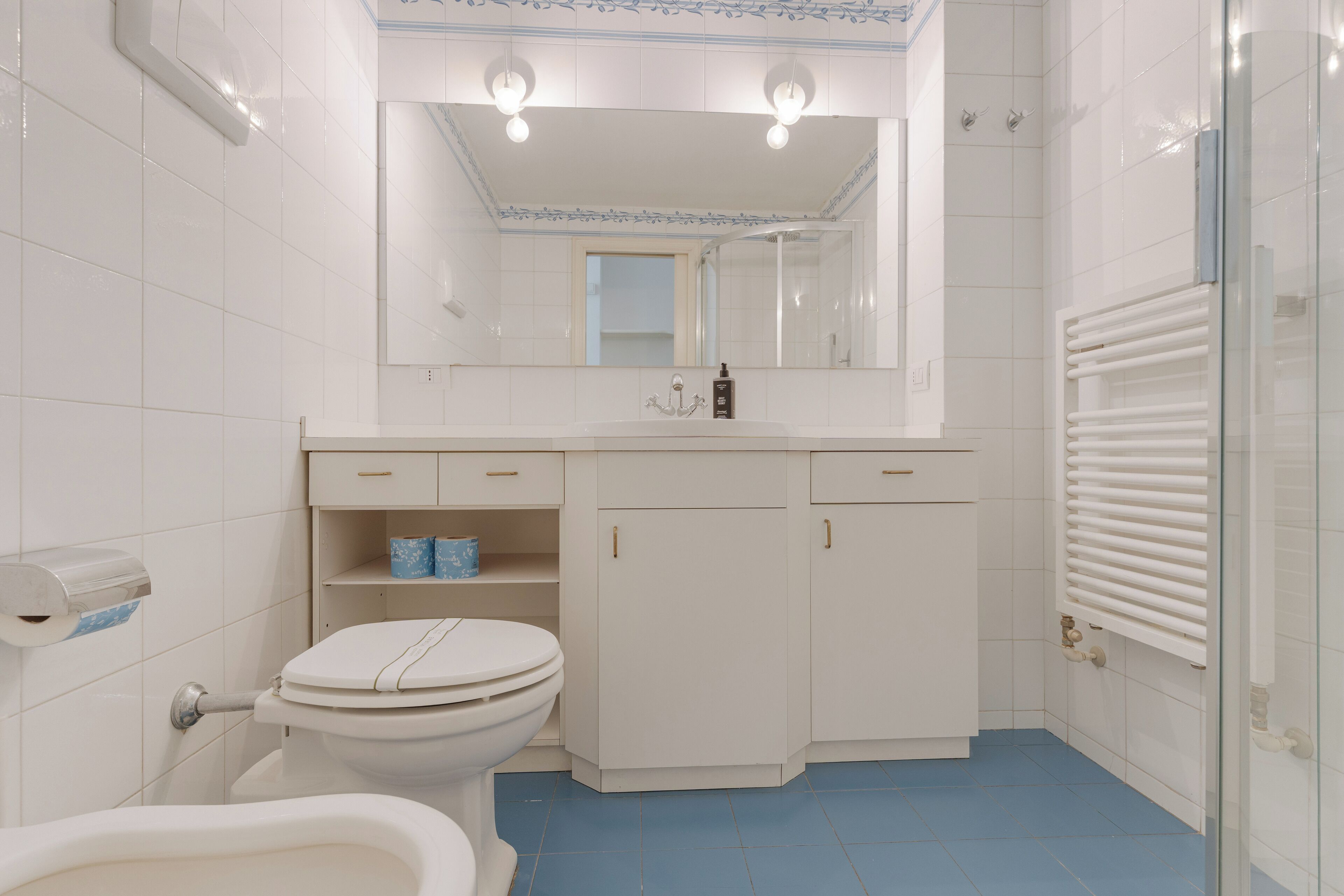 A bathroom with a toilet, a bidet, a sink with a mirror above, and a cabinet with a towel rack.