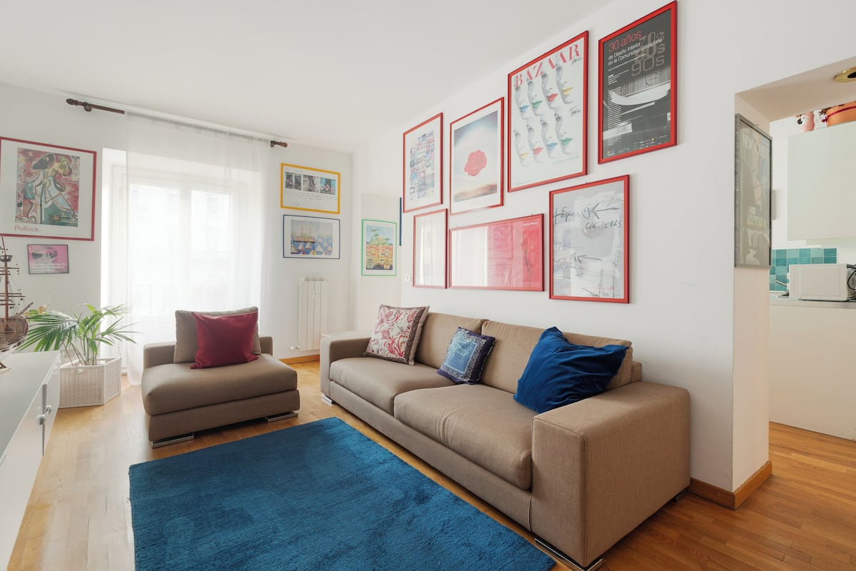 A living room with a sofa, a coffee table, and a blue rug. There are framed pictures on the wall and a window with curtains.