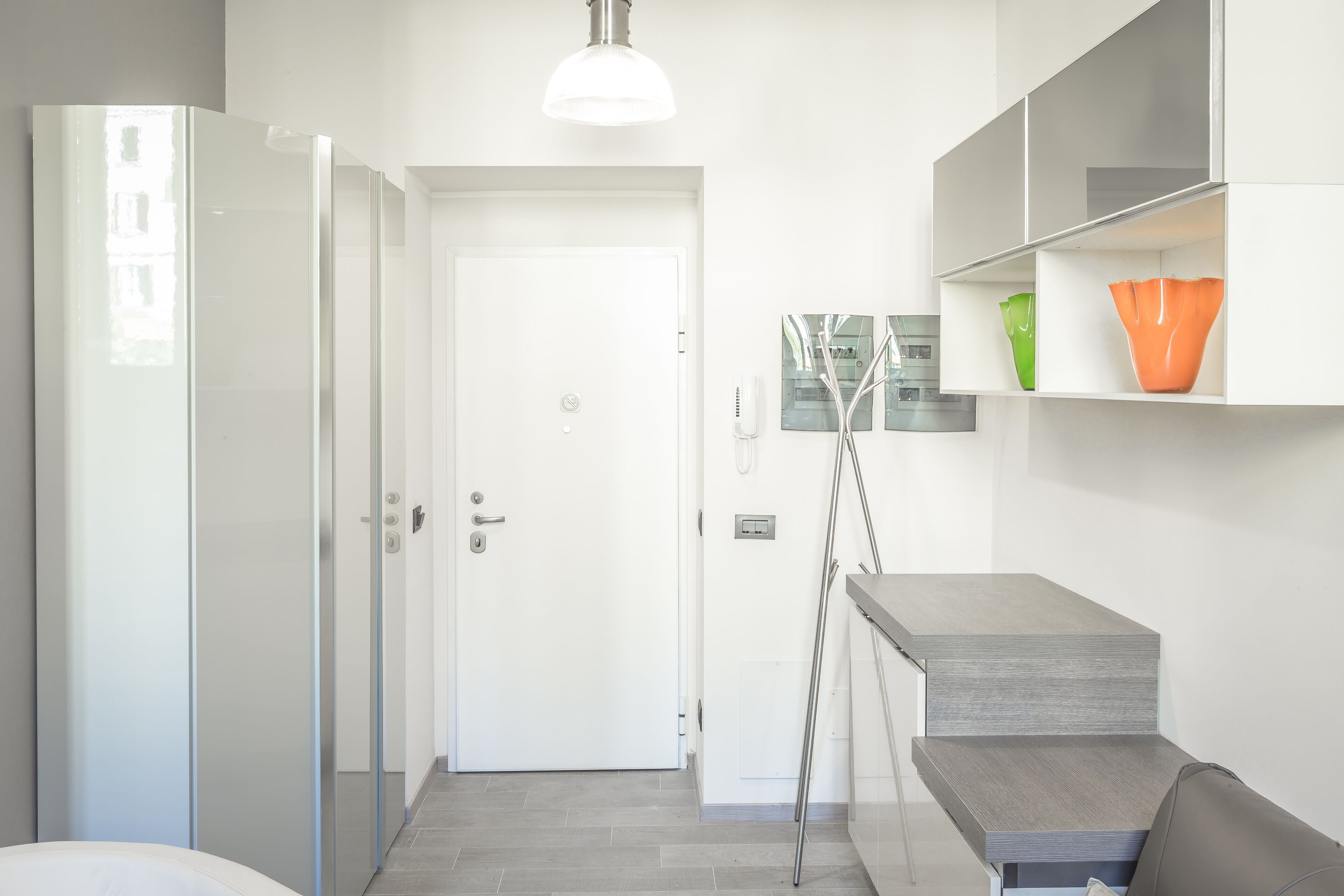 A modern hotel room with a glass door, a white wardrobe, a grey bench, and a small table with a lamp.