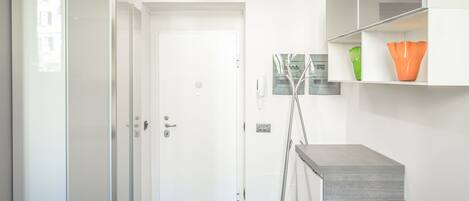 A modern hotel room with a glass door, a white wardrobe, a grey bench, and a small table with a lamp.