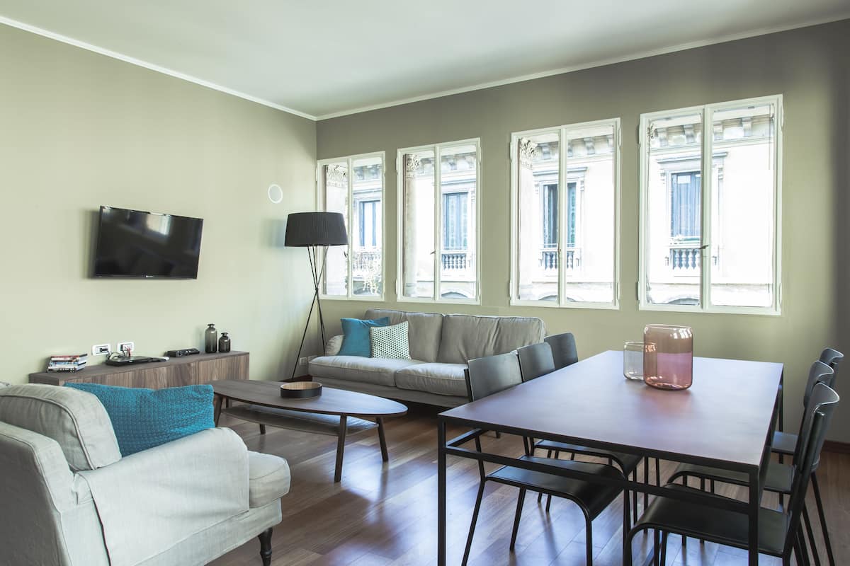 A modern living room with a sofa, coffee table, dining table, and a TV mounted on the wall.