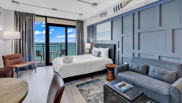 A hotel room with a bed, a grey sofa, a wooden coffee table, a desk, and a chair. There is a balcony with a view of the sky and clouds.