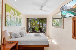 Une chambre à coucher comprenant un lit, une table de chevet, une télécommande, un ventilateur de plafond et une grande fenêtre donnant sur des arbres.