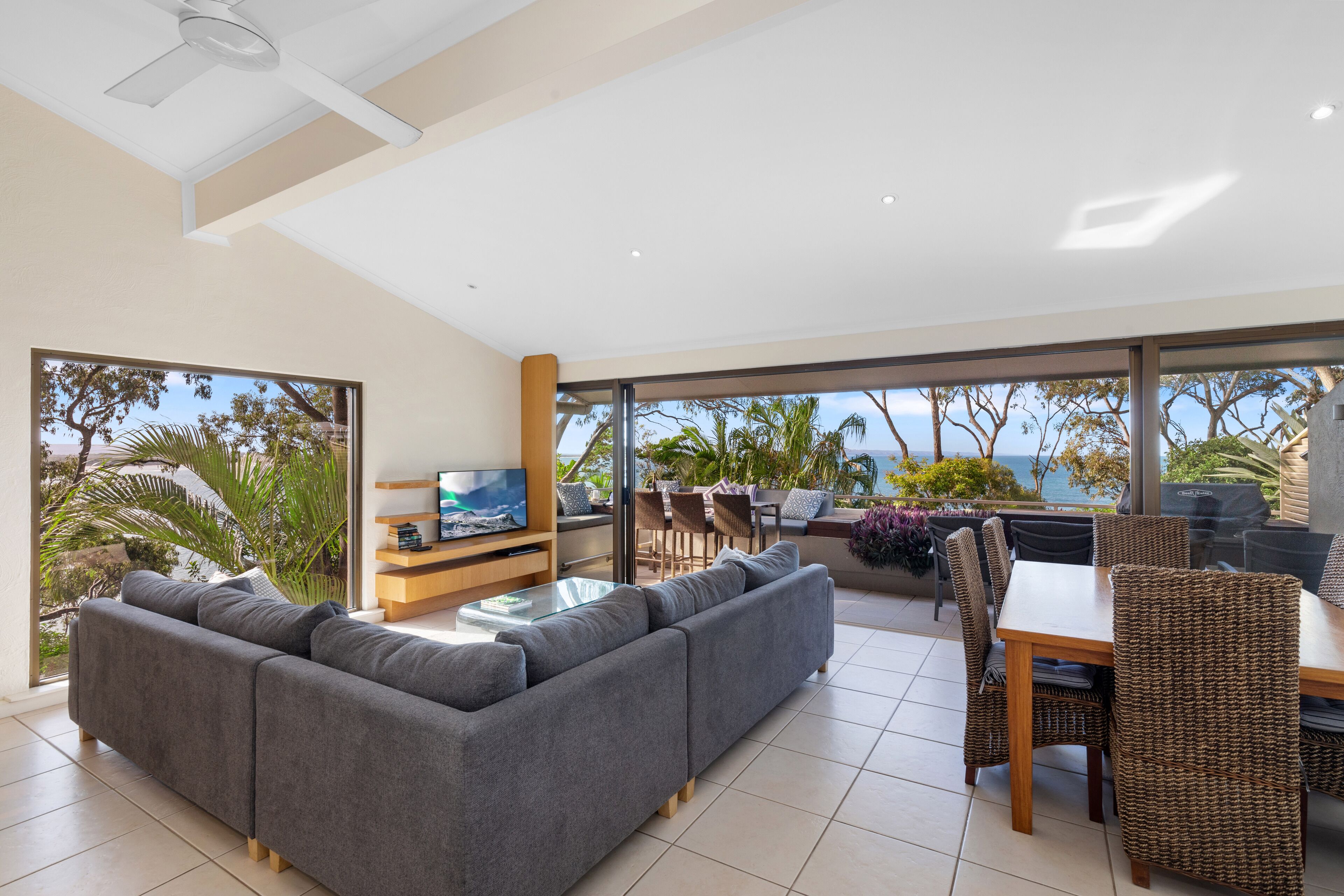 A modern living room with a grey sectional sofa, a glass coffee table, and a TV. There is a dining area with wicker chairs and a table, and large windows offering a view of the ocean and palm trees.