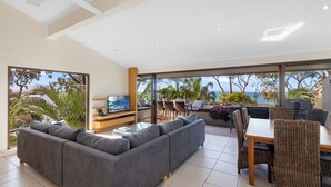 Una sala moderna con un sofá seccional gris, una mesa de centro de vidrio y una televisión. Hay una zona de comedor con sillas de mimbre y una mesa, y ventanas amplias que ofrecen vistas al océano y palmeras.