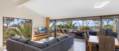 A modern living room with a grey sectional sofa, a glass coffee table, and a TV. There is a dining area with wicker chairs and a table, and large windows offering a view of the ocean and palm trees.