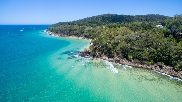 Vista aérea de una zona costera con aguas turquesas cristalinas, una playa arenosa y colinas boscosas de intenso verde.