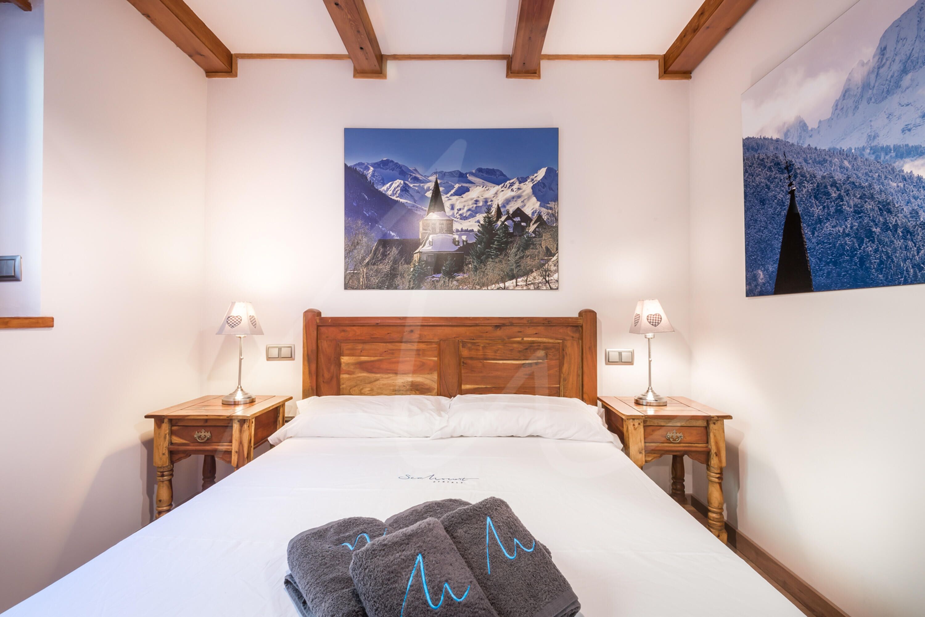 Une chambre d’hôtel avec un lit en bois, deux tables de chevet avec des lampes, un grand tableau représentant un paysage de montagne au-dessus du lit et un autre tableau représentant un paysage de montagne sur le mur.