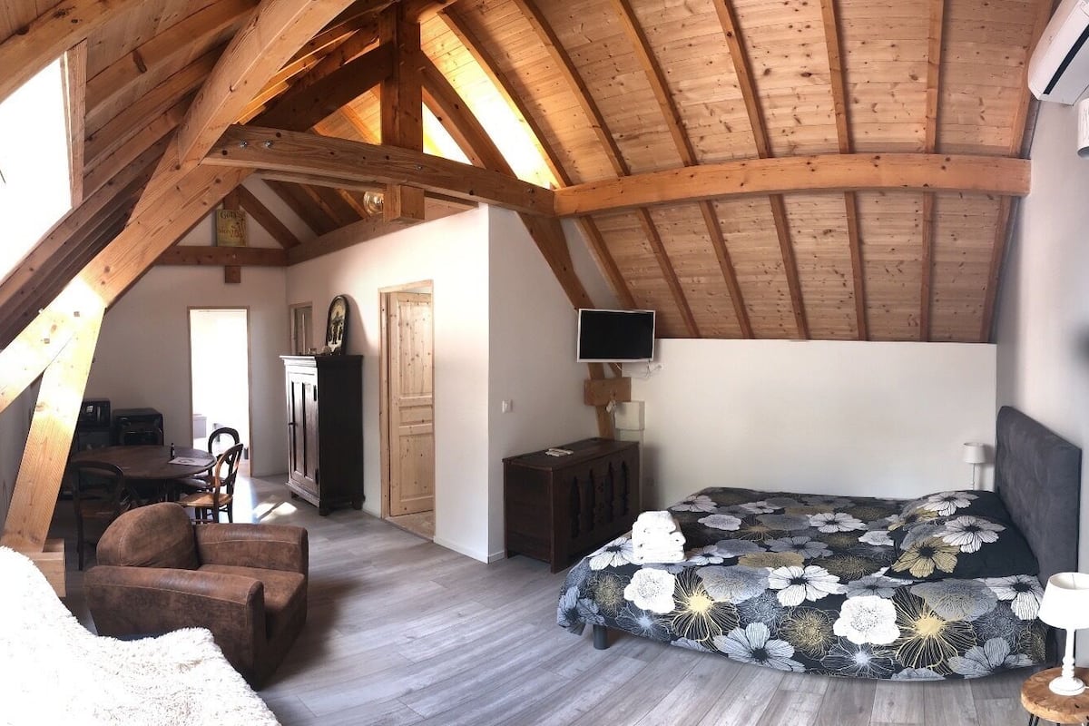 Une chambre avec un plafond en bois, un lit recouvert d’une couvre-lit à motifs floraux, un téléviseur fixé au mur et une chaise.