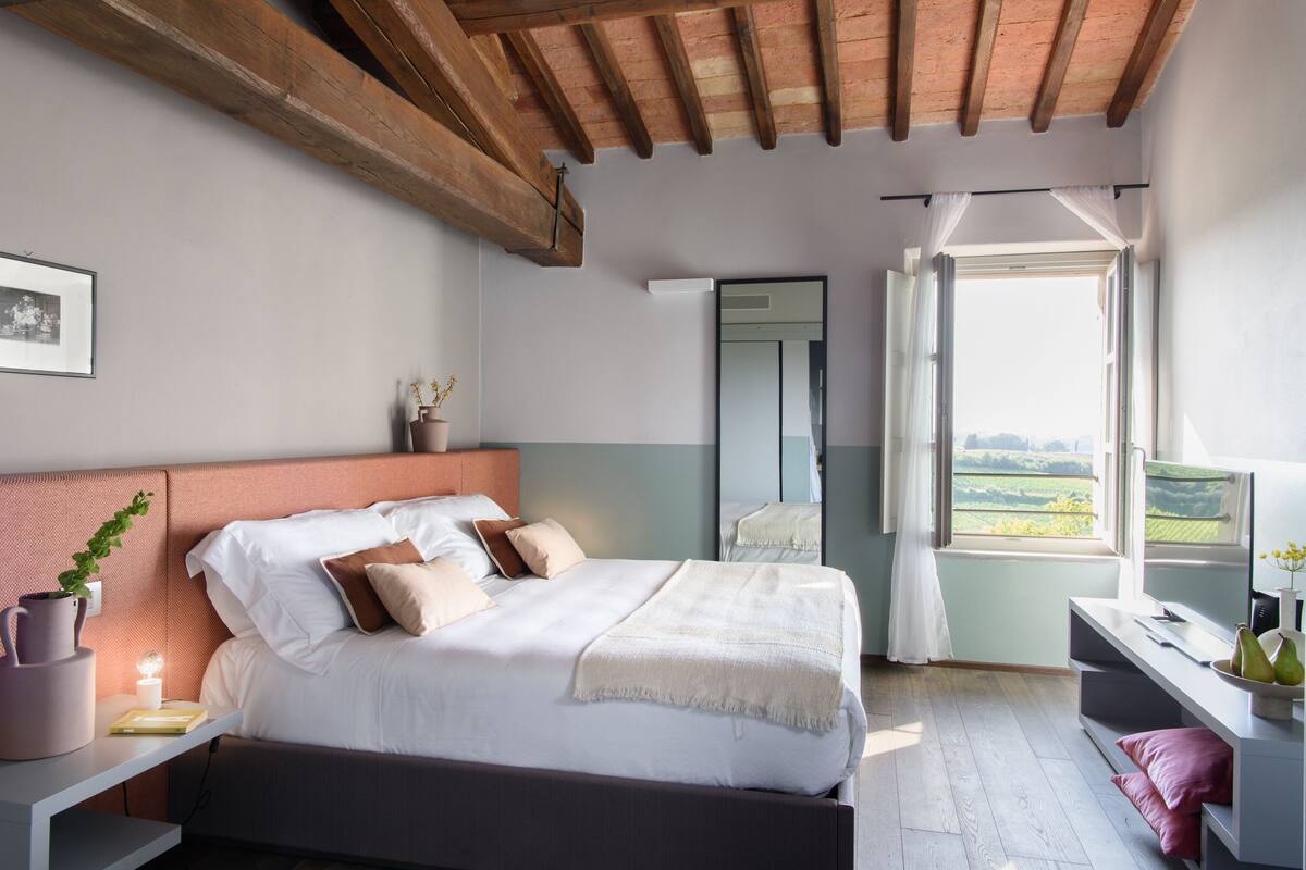 A bedroom with a large bed, a bedside table, a mirror, and a window with a view of the countryside.