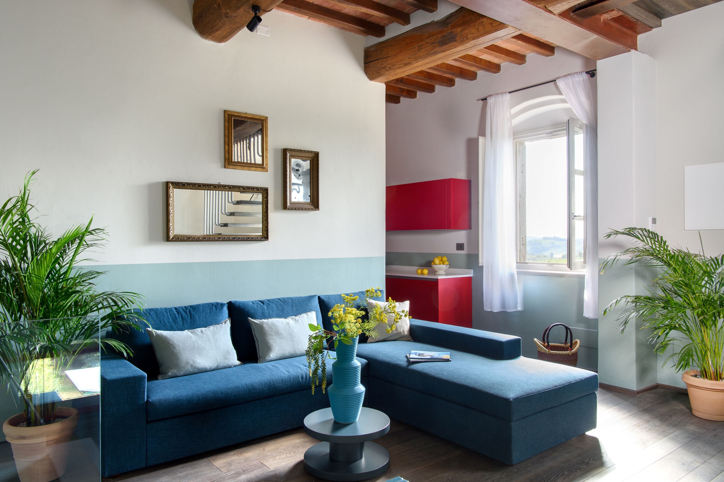 A living room with a blue sofa, a round coffee table with a vase, and a view of a red kitchen through an open door.