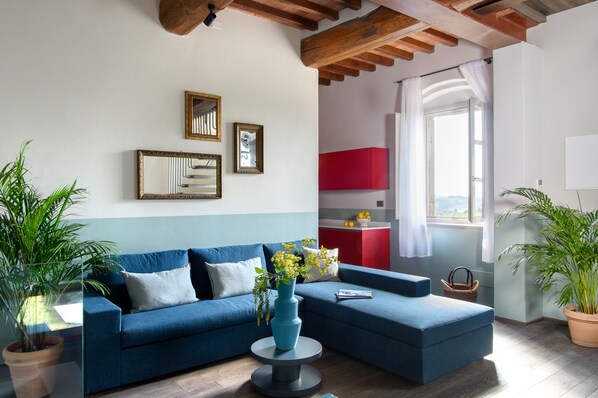 A living room with a blue sofa, a round coffee table with a vase, and a view of a red kitchen through an open door. - Laqua Vineyard (Terricciola)