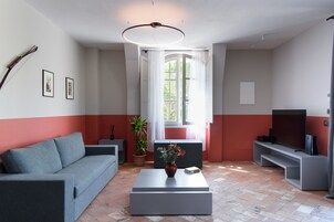 Uma sala de estar moderna com um sofá cinzento, uma mesa de centro com um vaso, uma televisão sobre um móvel e uma janela grande com cortinas.