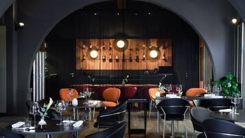 Un restaurante moderno con interior oscuro, sillas rojas y negras, y una pared decorada con botellas colgantes.