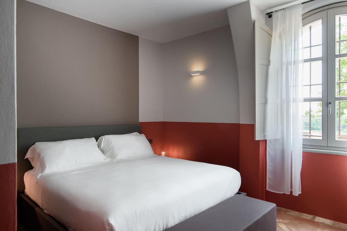 A bedroom with a bed, a nightstand, a wall-mounted light, and a window with curtains.