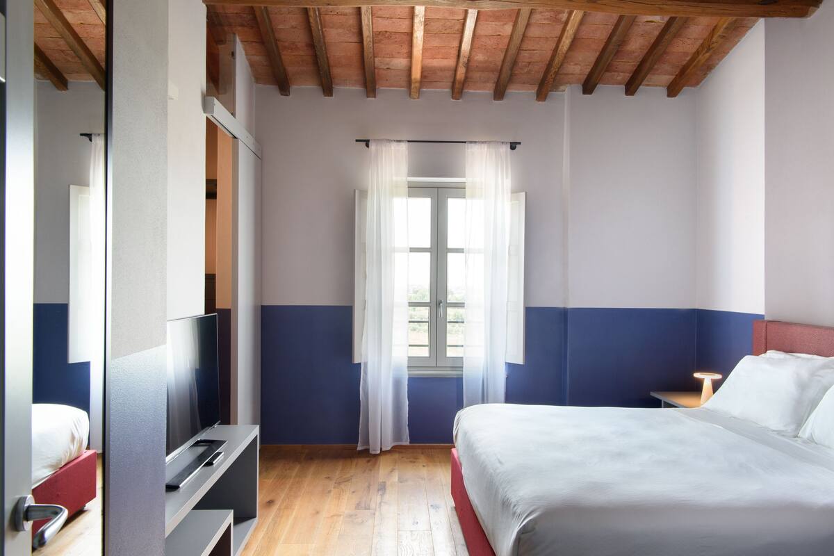 A hotel room with a bed, a TV, a window with curtains, and a wooden ceiling.