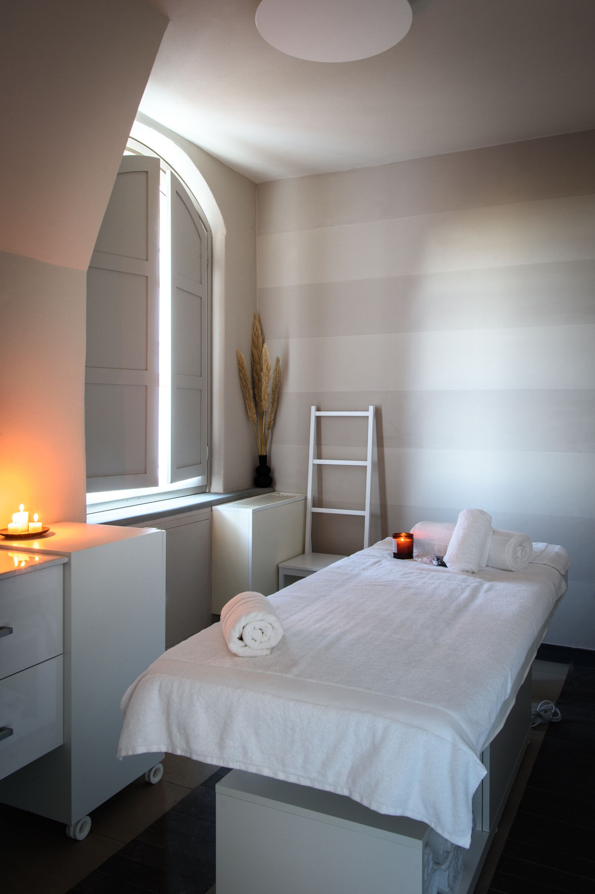 Un dormitorio con una cama, una ventana, una mesita de noche con una vela, un radiador y un aparador blanco.