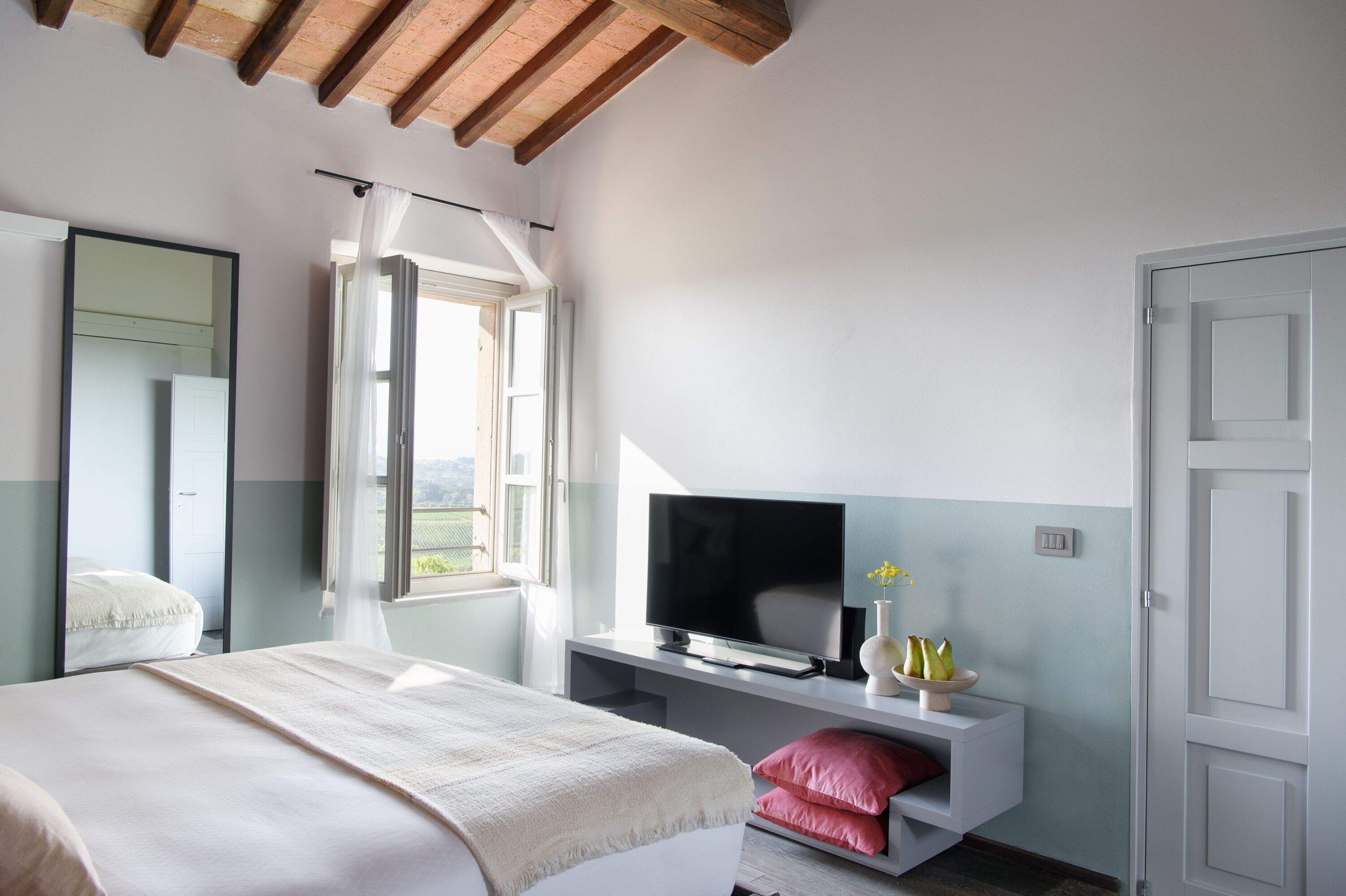 Un dormitorio con cama, televisión sobre un mueble, ventana con cortinas y puerta.
