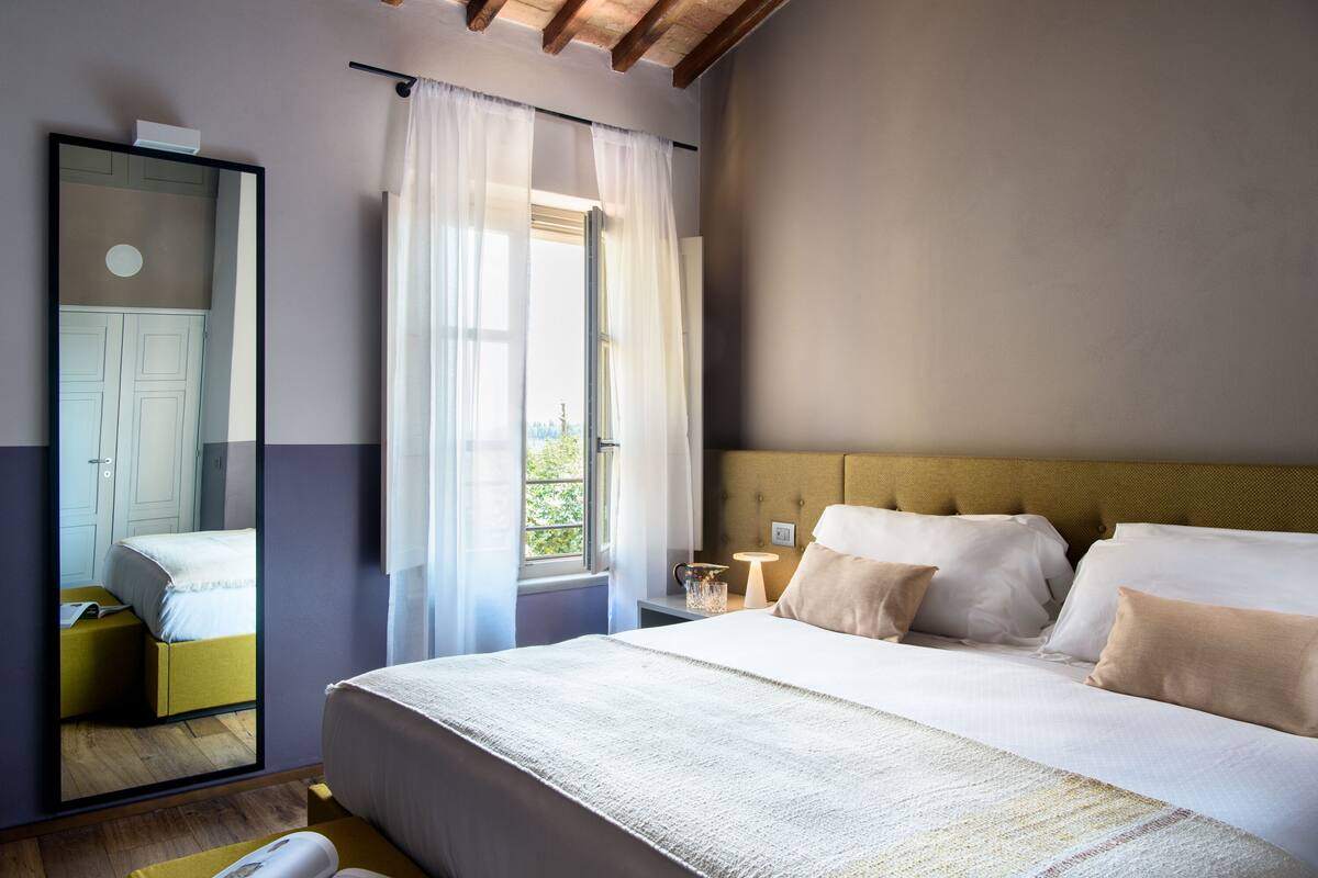 A bedroom with a large bed, a bedside table with a lamp, a mirror, and a window with curtains.