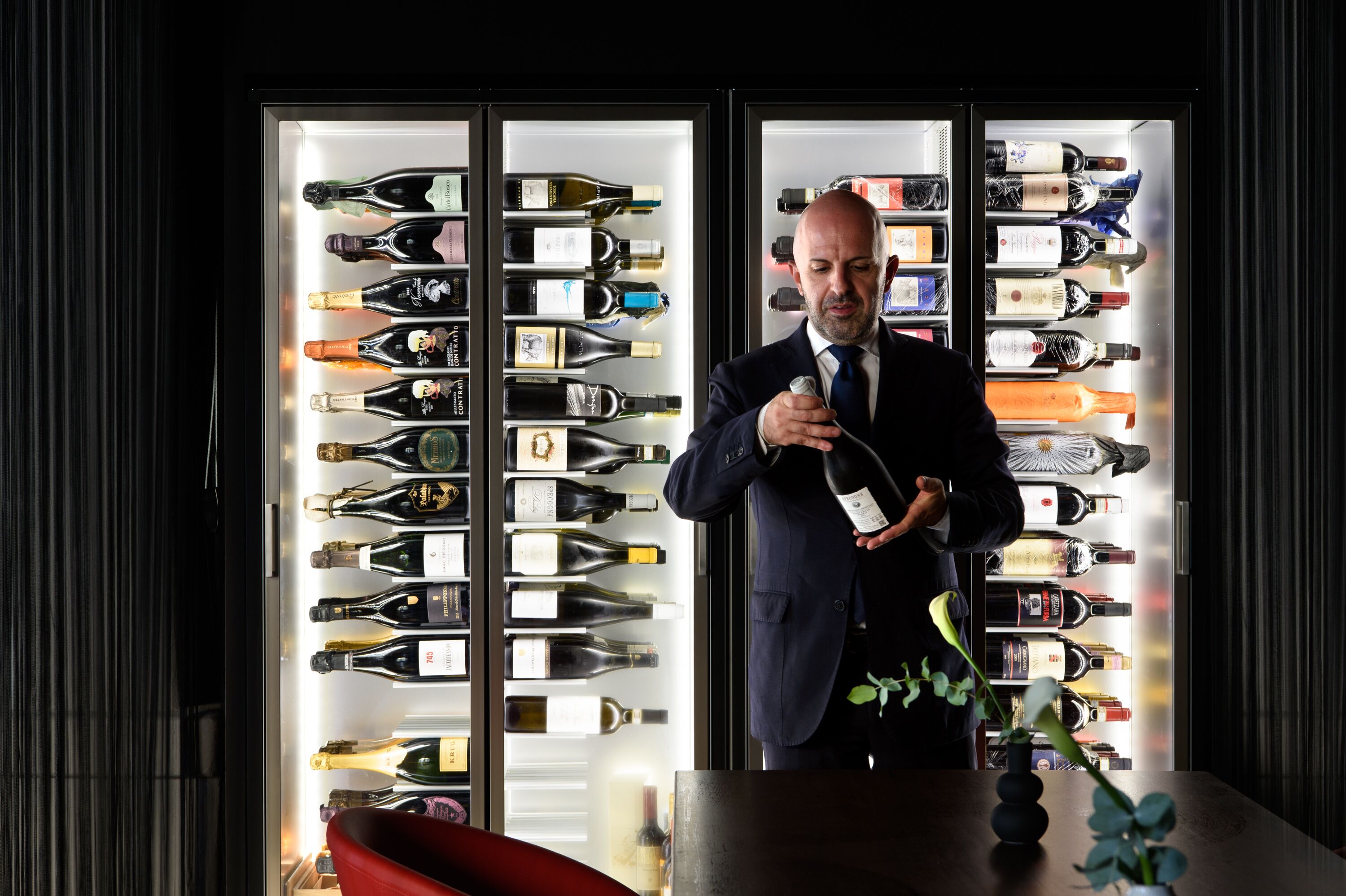 A man in a suit holding a wine bottle in front of a wine refrigerator filled with various bottles.