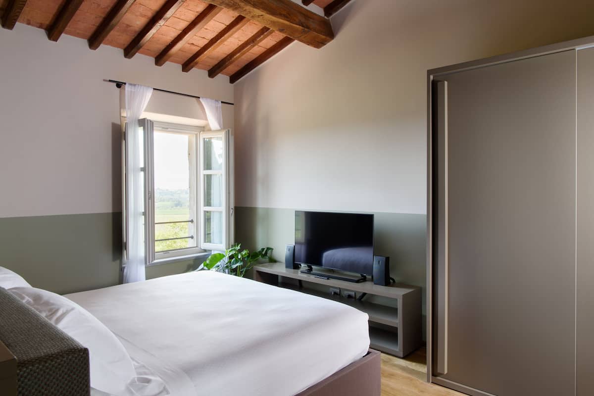 A bedroom with a bed, a TV on a stand, a window with curtains, and a door.