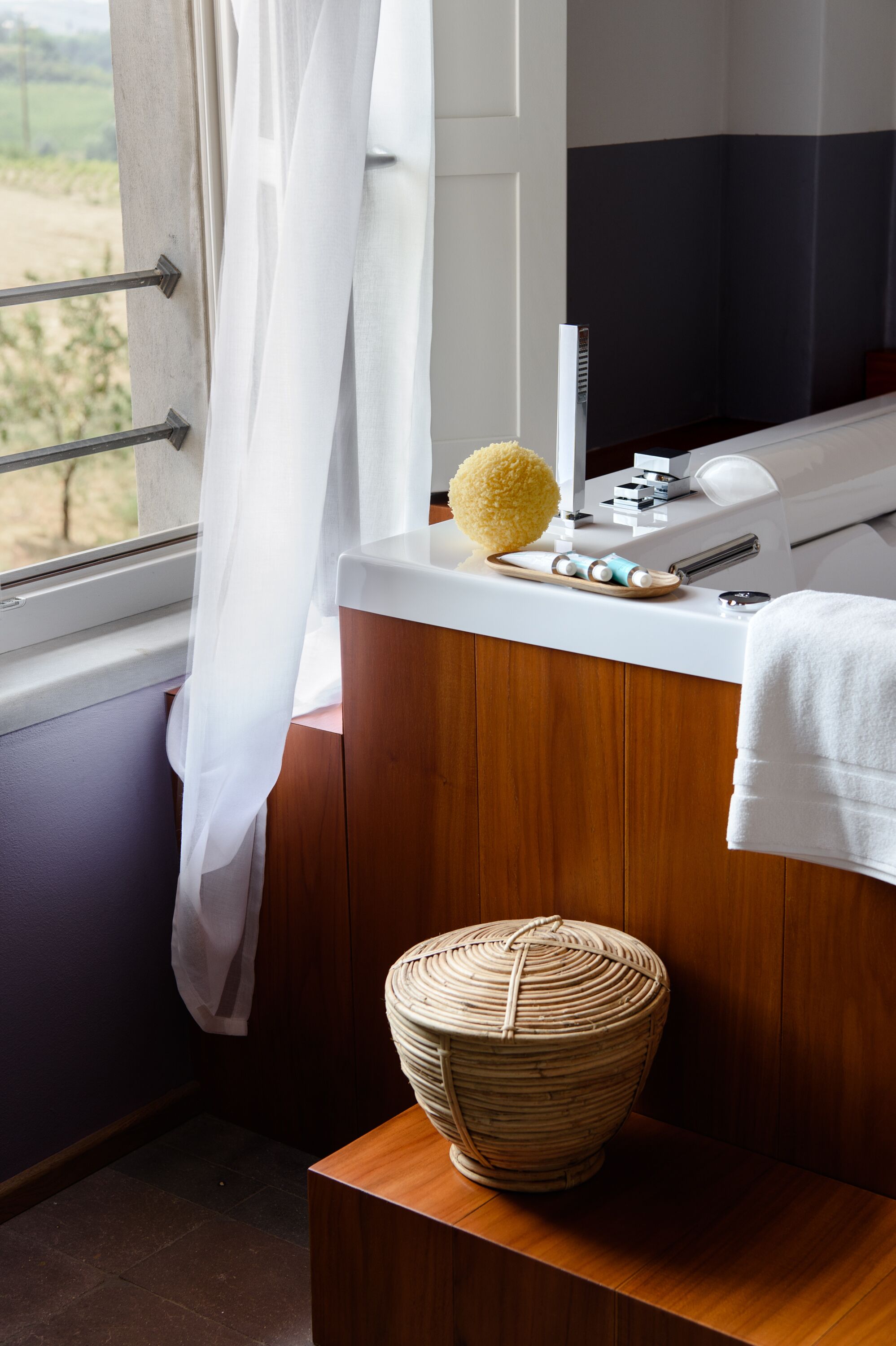 A bathroom with a wooden vanity, a white sink, a yellow bath sponge, a woven basket, and a window with a view of greenery.