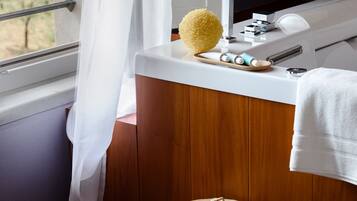 A bathroom with a wooden vanity, a white sink, a yellow bath sponge, a woven basket, and a window with a view of greenery.