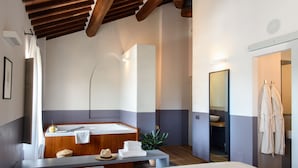 Una habitación de hotel moderna con una cama grande, un banco de madera, una mesita con toallas y un sombrero, una planta y un baño visible a través de una puerta abierta.