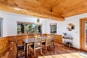 Zona pranzo con tavolo e sedie in legno, ampie finestre con tende e lampadario a sospensione.