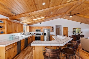 A kitchen with wooden cabinets, stainless steel appliances, and a large island with bar stools. - Sky Pine Retreat by Destination Big Bear: Log Cabin Home! Game Room! HOT TUB! EV Charger! (Big Bear Lake)