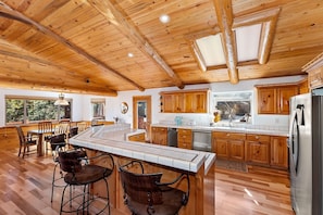 A spacious kitchen with wooden cabinets, a large island with bar stools, and a view of the outdoors through a window. - Sky Pine Retreat by Destination Big Bear: Log Cabin Home! Game Room! HOT TUB! EV Charger! (Big Bear Lake)
