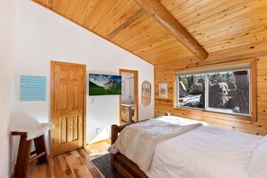 Una habitación con paneles de madera, una cama, un taburete de madera, una puerta y una ventana con vista a la montaña.