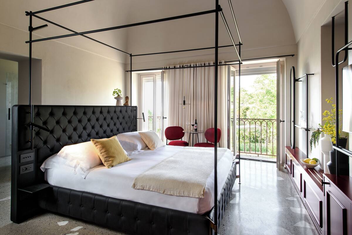 A four-poster bed with a tufted headboard, a bedside table, and a view of a balcony with red chairs and a table.