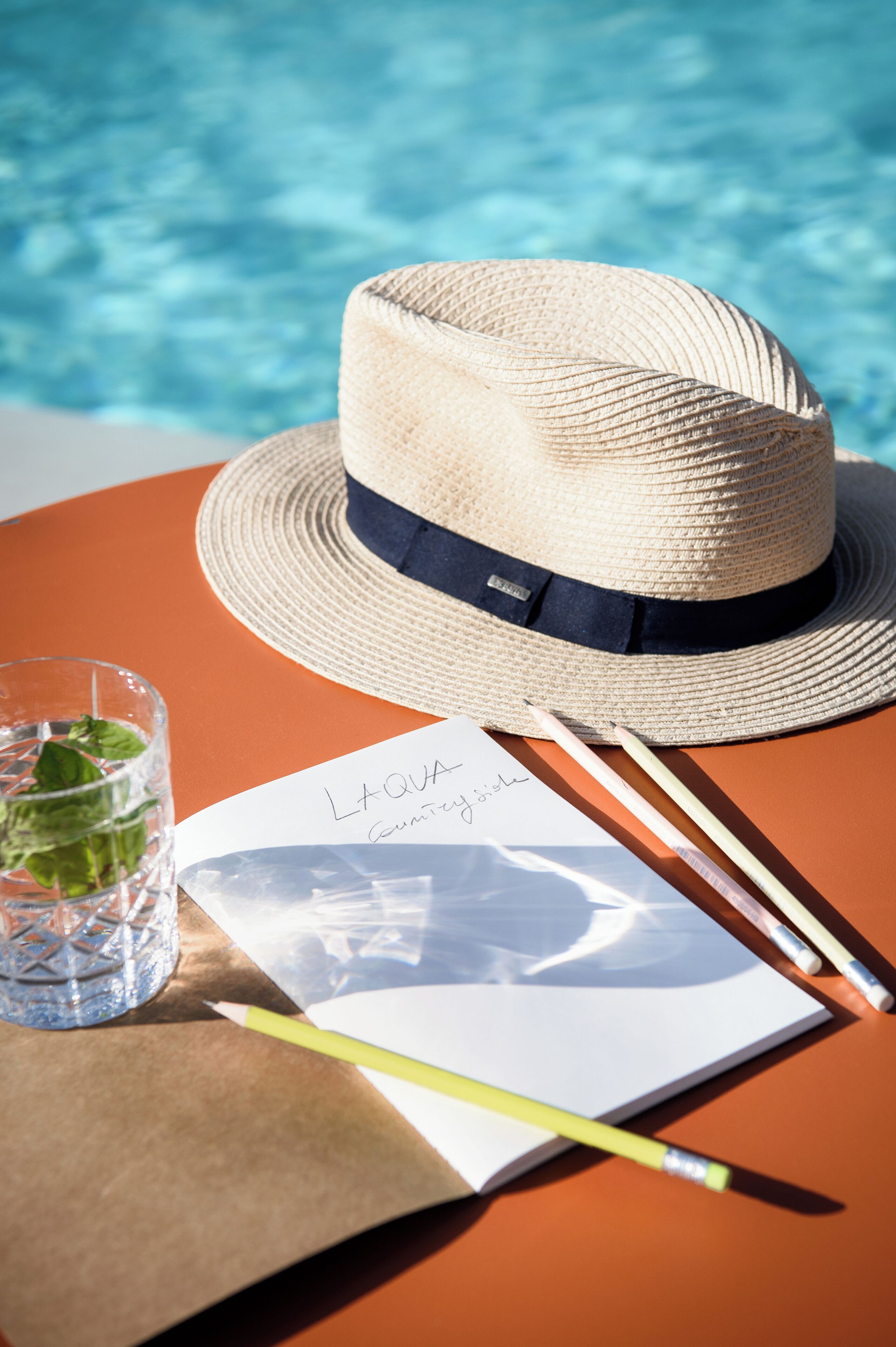 A straw hat, a glass of water, a notebook, and pencils on a table by a pool.