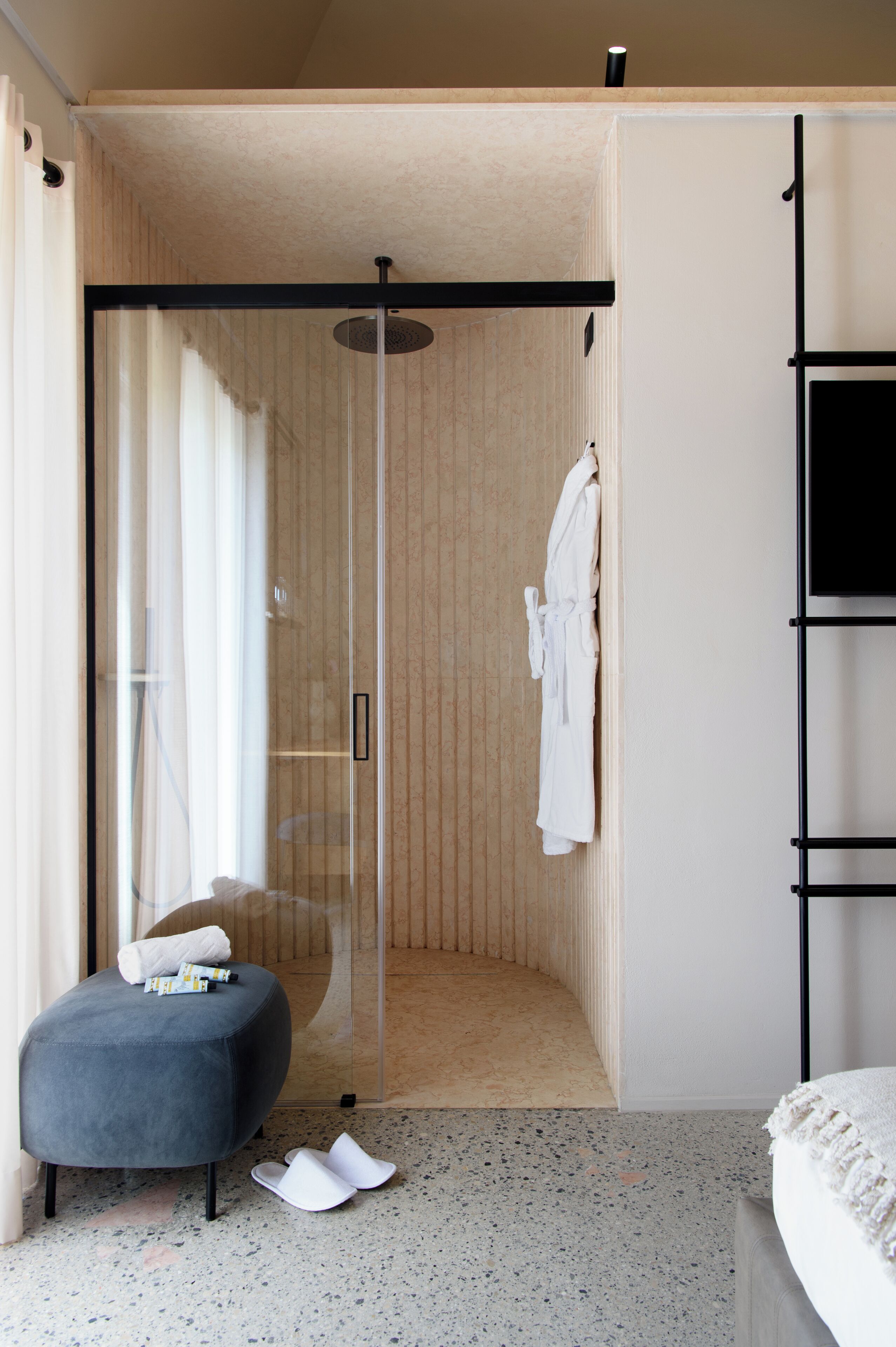 A modern bathroom with a glass shower enclosure, a towel rack, and a small seating area with slippers.