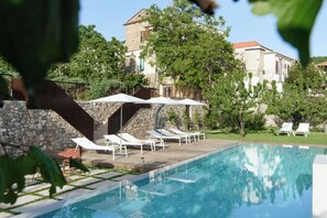 Piscina con sillas de descanso y sombrillas, rodeada de muros de piedra y vegetación.