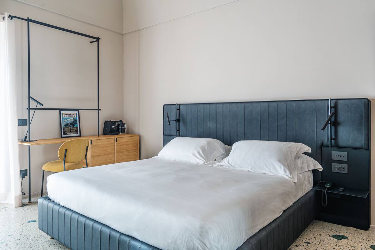 A neatly made bed with white linens, a wooden desk with a chair, and a nightstand with a phone and a lamp.