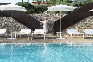 Zona de piscina con sillones de lounge blancos y sombrillas, una pared de piedra y una persona con bata blanca.
